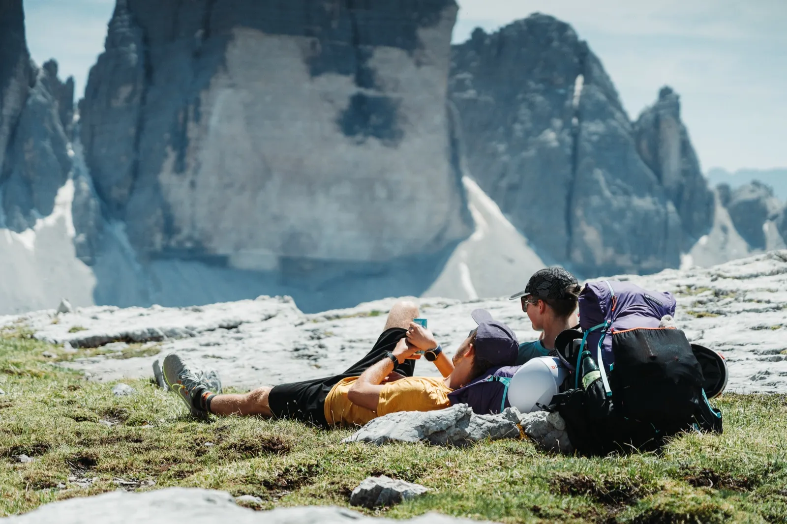Two hikers lie on the grass resting with a view of the Tre Cime