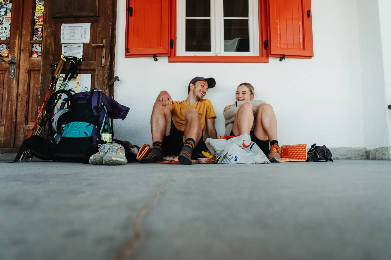 A couple sit with their backpacks against a wall, eating lunch in the shade of the hut