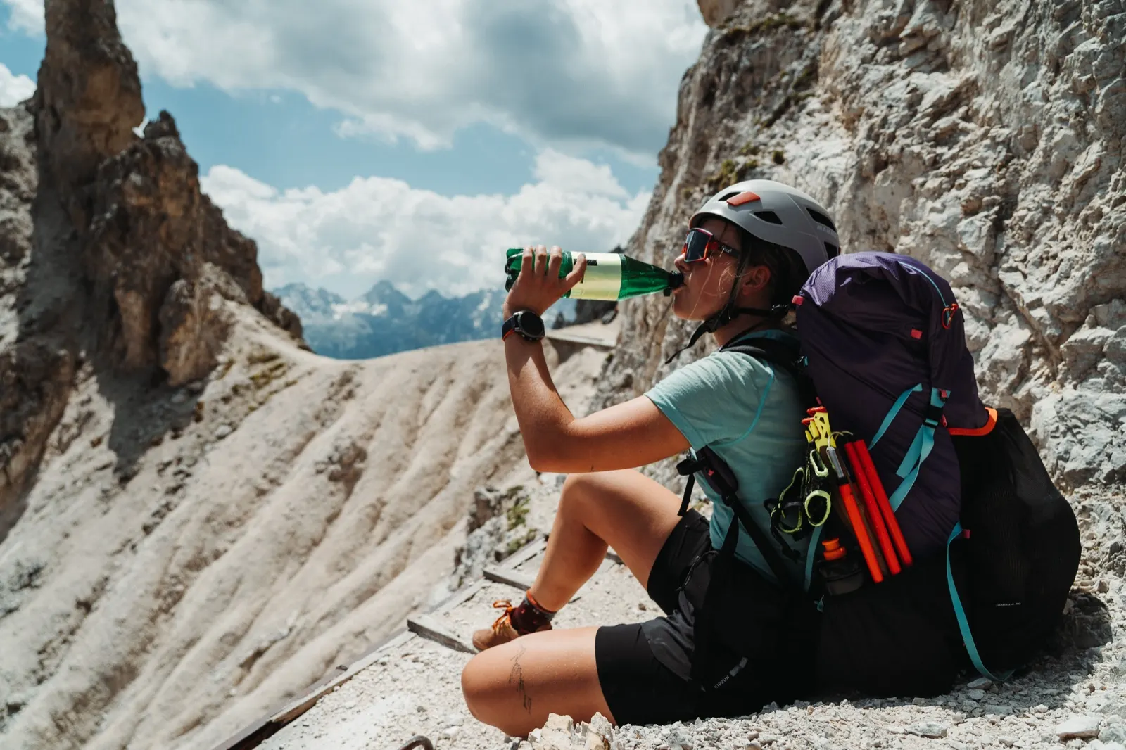 Hiker in a helmet sits on a rock drinking water during a break in the mountains