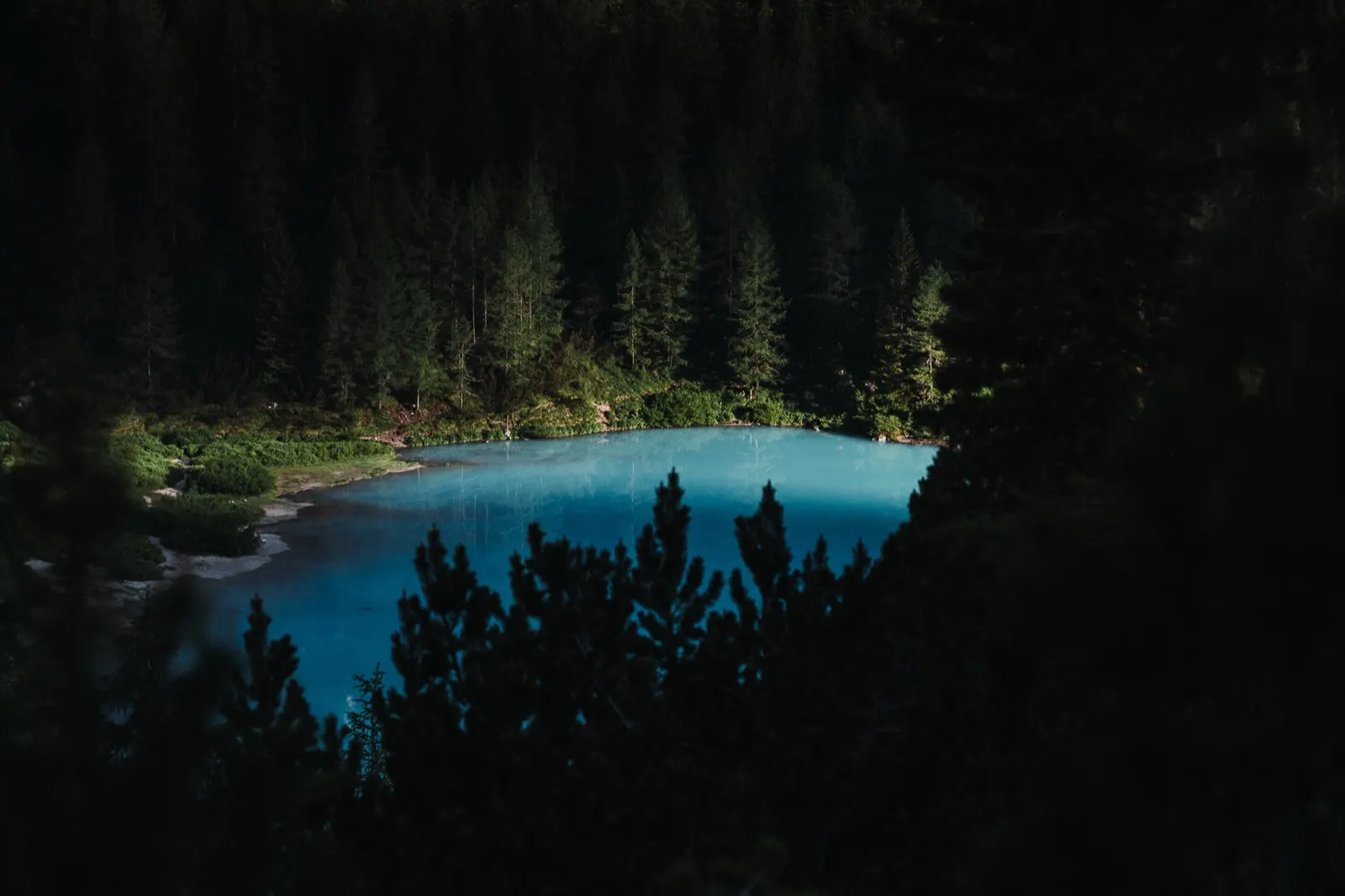 A view of a turquoise lake surrounded by dark forest, lit by a soft beam of falling light.