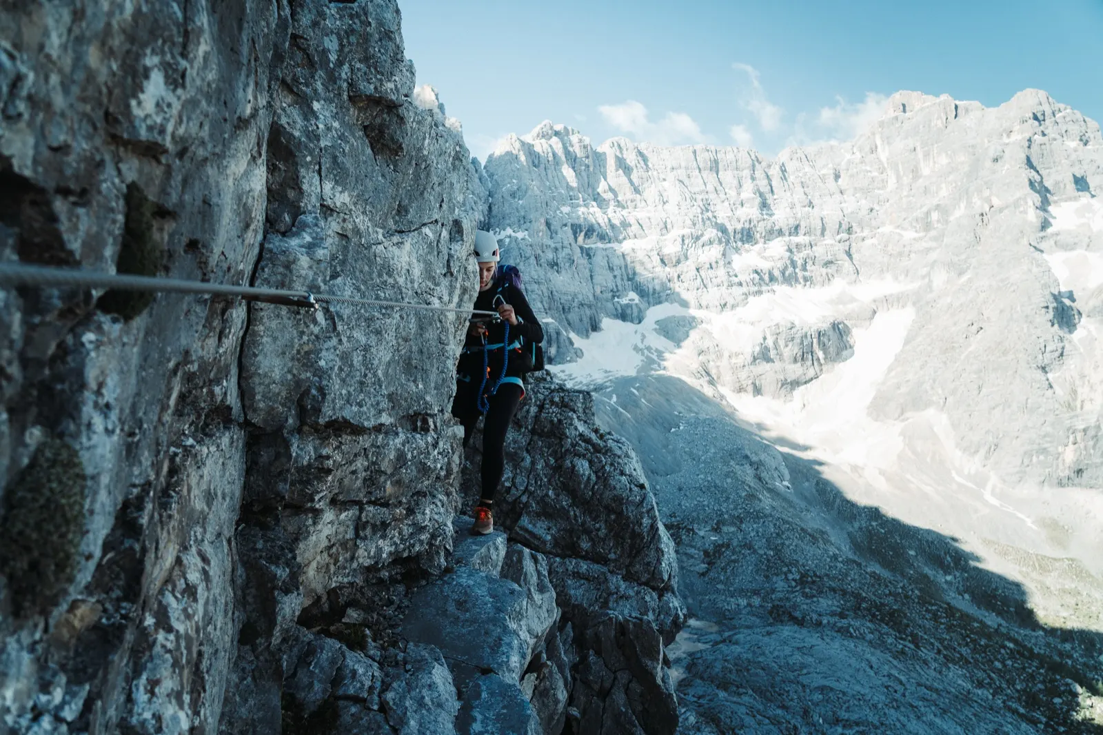 Hiker with gear walking along a narrow secured path on a rock face with a view of the peaks.
