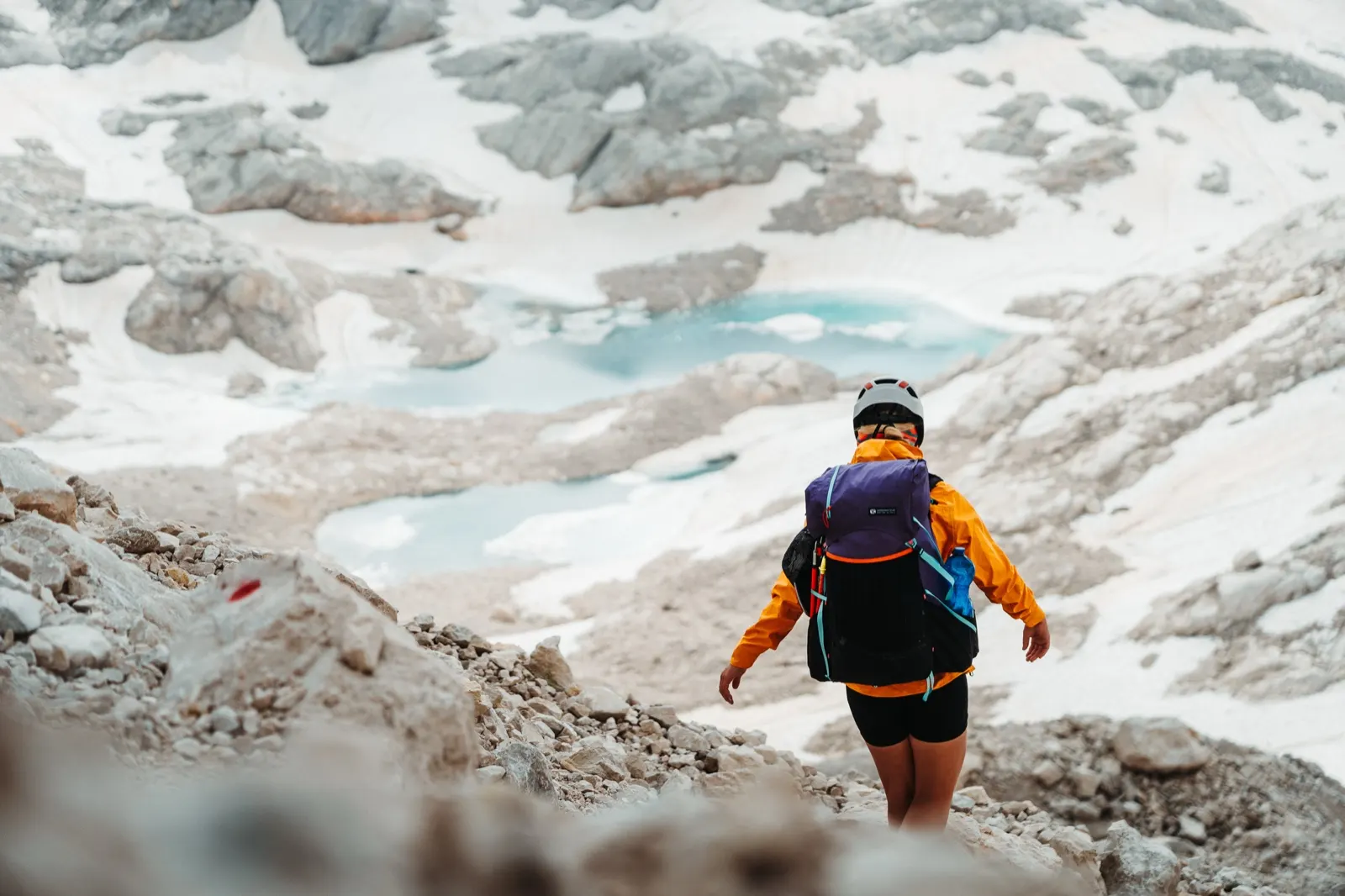 A view of a hiker descending to turquoise lakes between snowfields.