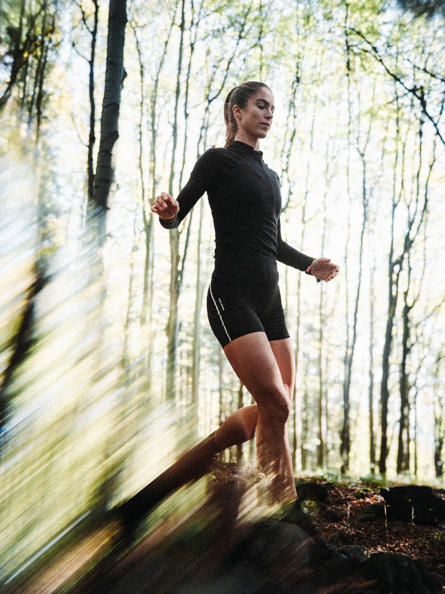 Runner in a black long-sleeve top and shorts leaping over a rock in a bright beech forest, shot from a low angle