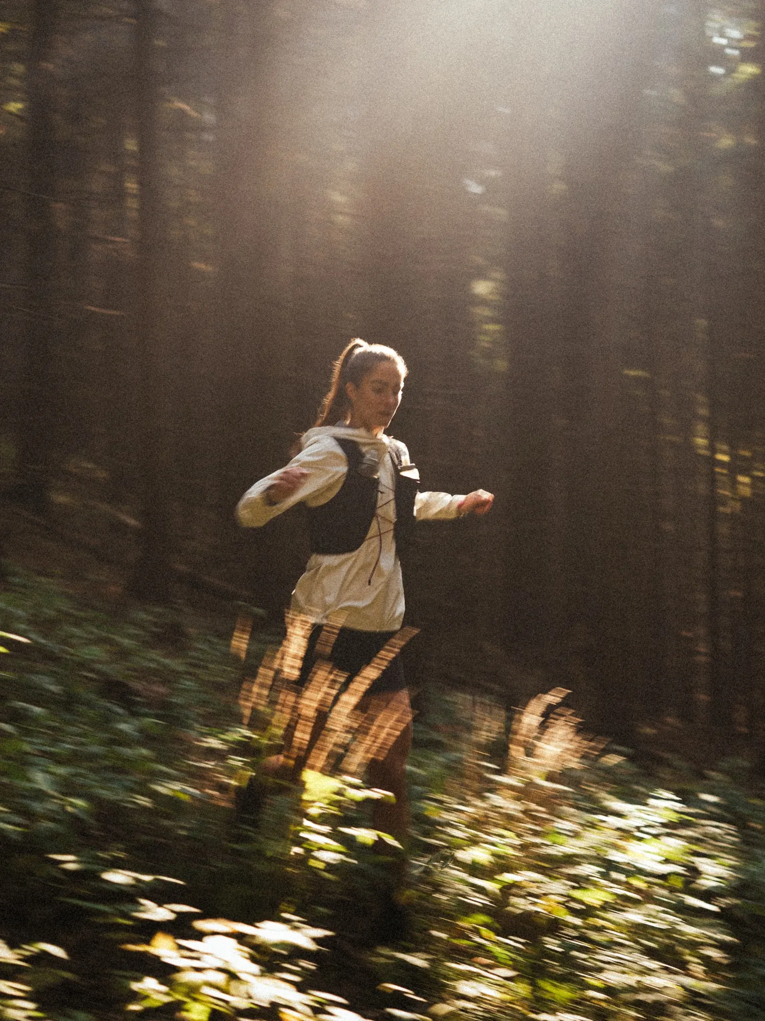 Runner in a cream jacket and vest in a dark spruce forest with backlit ferns in the foreground