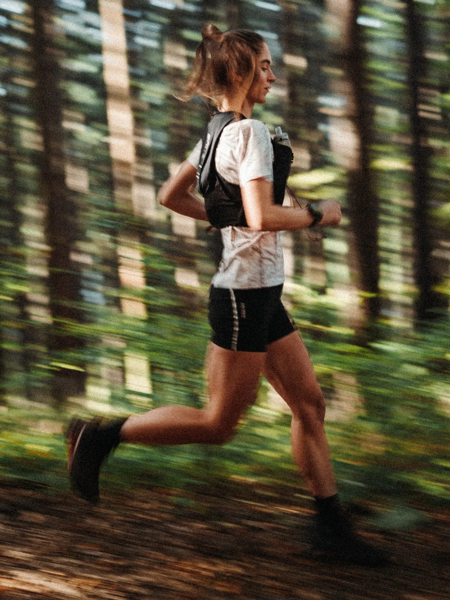 Side view of a runner in a white tee and running vest with a soft flask, trees blurred behind her