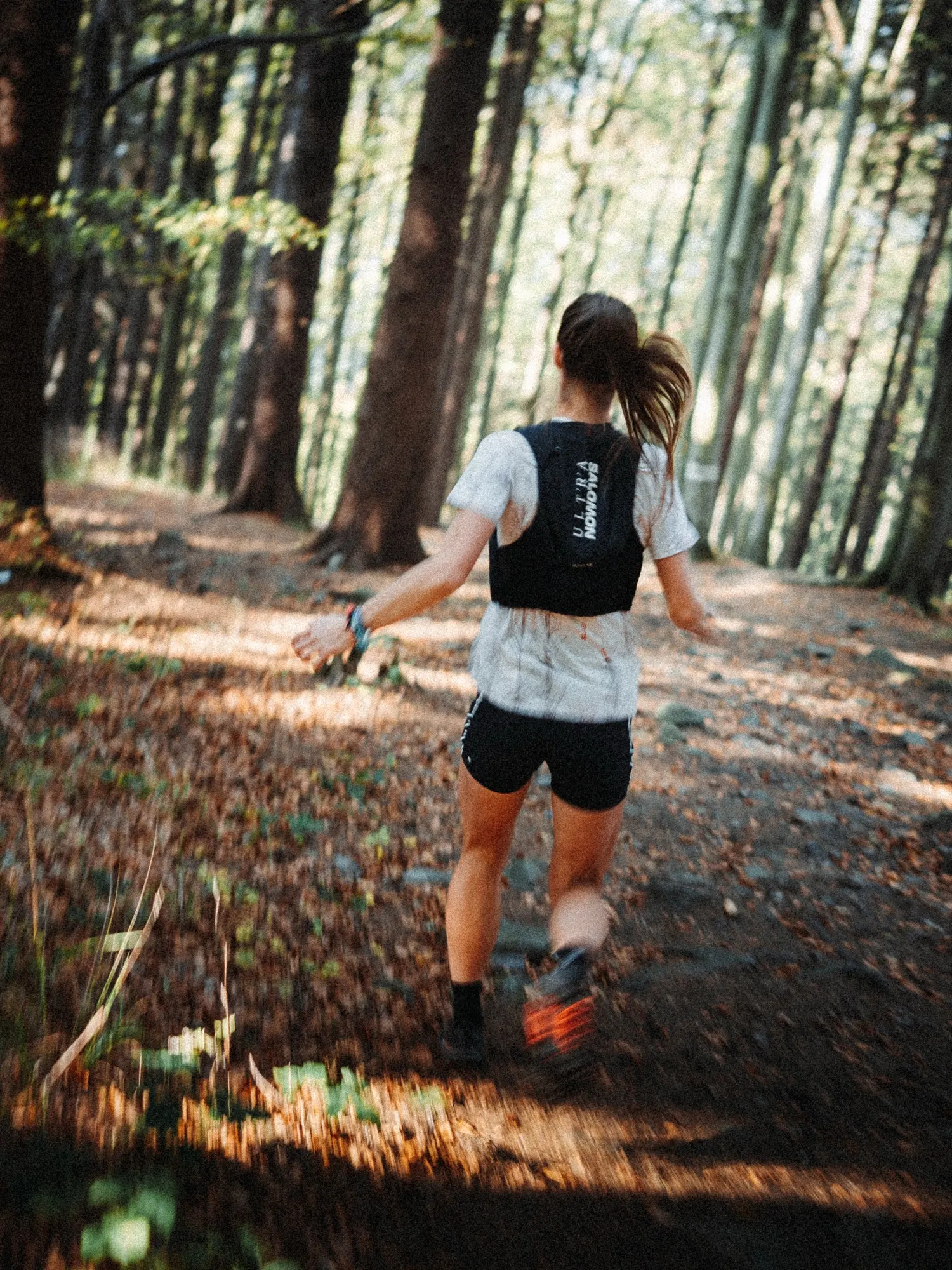 Runner seen from behind in a Salomon Ultra running vest on a sunlit forest trail