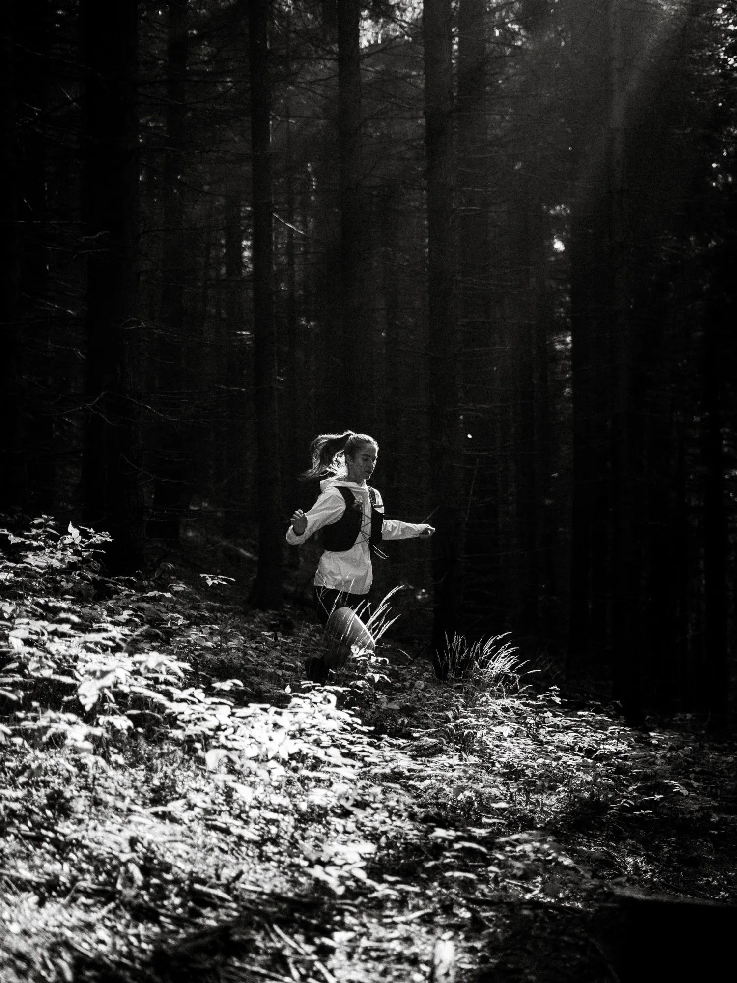 Black-and-white shot of a runner in a light jacket and vest bounding through undergrowth in a dark forest