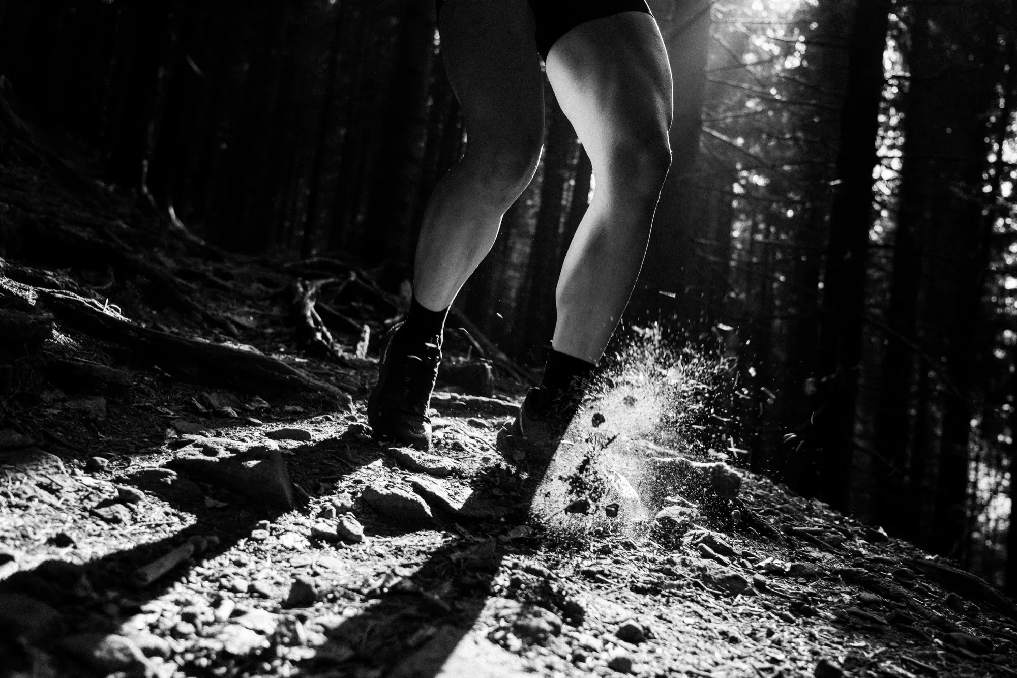 Black-and-white close-up of a trail runner's legs and shoes kicking up dust on a rocky forest path