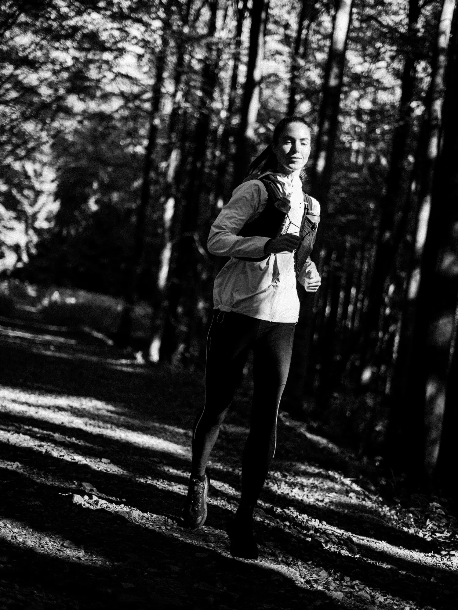 Black-and-white portrait of a runner in a light jacket and vest on a sunlit trail among tall trees