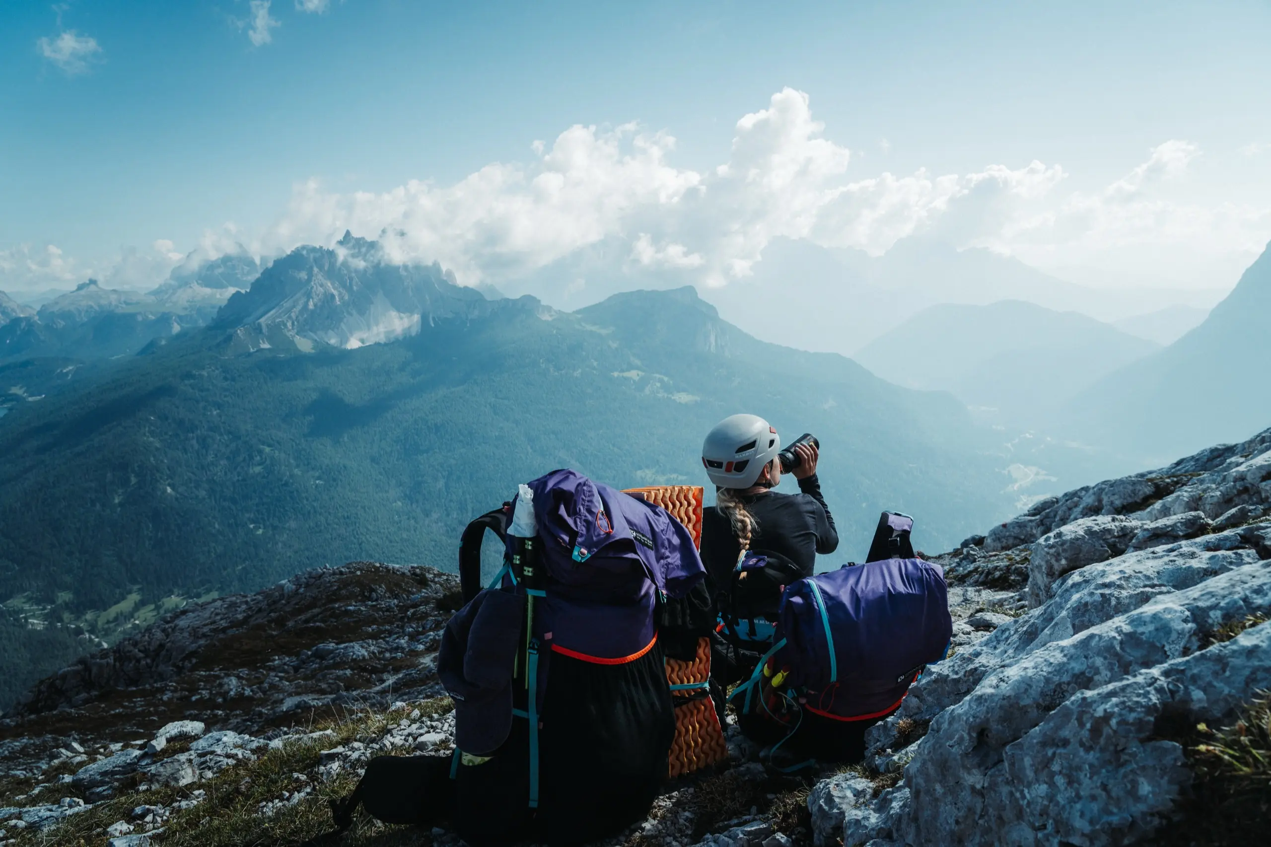 Turista v helmě odpočívá u dvou batohů na skalnatém hřebeni nad dolomitským údolím